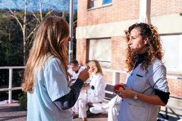 Two healthcare workers engaging in a conversation while holding fresh apples on a sunny break outside the hospital building