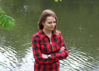 A young girl in a red and black dress walks against the background of a pond in the city in summer