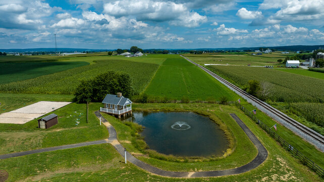 A Small Pond With A Water Fountain Is Situated Near A Curved Pathway And A Quaint House, With Parallel Railroad Tracks And Expansive Crop Fields Stretching Into The Distance Beneath A Cloudy Sky.