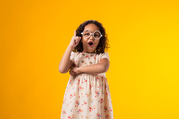 Surprised kid girl with glasses looking at camera over yellow background. Childhood and knowledge concept