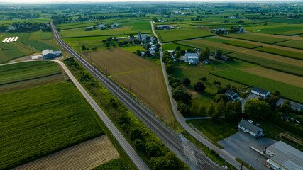 Aerial View Of A Curved Road Running Parallel To Railroad Tracks, Flanked By Farmhouses And Fields, In A Verdant Agricultural Landscape During Early Evening.