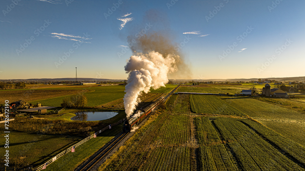 Wall mural An Aerial View of Historic Steam Train Emitting A Massive Plume Of Smoke As It Travels Through A Lush Countryside With Farm Buildings At Sunrise. - Wall murals