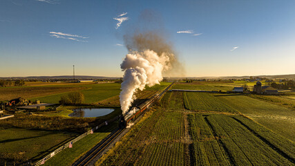 An Aerial View of Historic Steam Train Emitting A Massive Plume Of Smoke As It Travels Through A Lush Countryside With Farm Buildings At Sunrise.