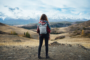 A traveler woman standing on a rugged hilltop, gazing at a distant mountain range with snowy peaks, under a dynamic sky, evoking a sense of adventure and exploration. 