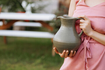 A woman in a pink dress holds a gray ceramic jug outdoors. Concept of handicraft ceramics or...