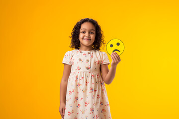 Smiling kid girl holding sad emoticon. Mental health, psychology and children's emotions concept.