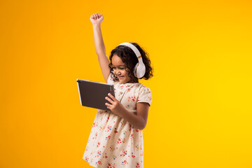 Smiling kid girl with headphones using tablet and shoeing winner gesture. Lifestyle, leasure and gadget addiction concept