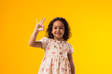 Smiling child girl showing ok gesture over yellow background. Positive emotions concept