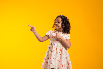 Smiling child girl pointing to an empty place on yellow background. Advertisement concept