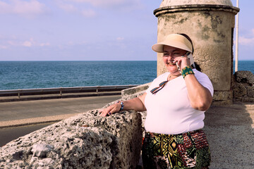 Medium shot of an older woman standing, leaning against the wall of the terrace in the city of Cartagena, wearing casual attire and a visor, talking cheerfully on the phone.