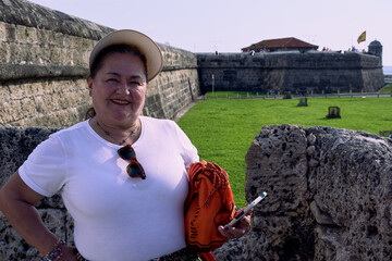 Portrait of an older woman from the waist up on the walls of Cartagena, dressed in a white blouse, wearing a visor on her head, and holding an orange Wayuu backpack and her phone in her left hand.