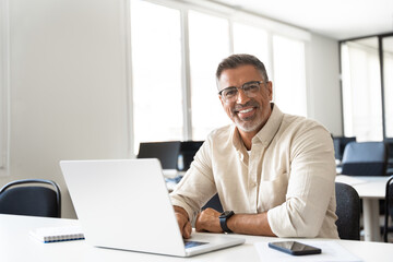 Fototapeta na wymiar Portrait of mature Indian or Latin business man ceo trader using laptop computer, typing, working in office. Middle-age Hispanic smiling handsome businessman entrepreneur looking at camera. Copy space