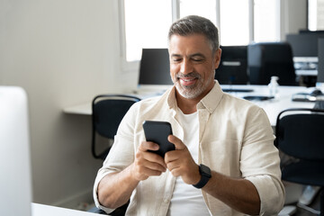 Middle aged Hispanic business manager ceo using cell phone mobile app, laptop. Smiling Latin or Indian mature man businessman holding smartphone sit in office working online on gadget with copy space.