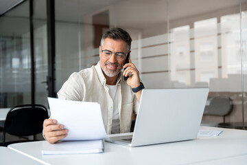 Middle aged Latin businessman having call on smartphone with business partners or clients. Smiling mature Hispanic man sitting at table talking by mobile cellphone, using laptop, paper documents