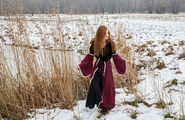 A red-haired girl with flowing hair in a beautiful burgundy dress on a spring snow-covered field.