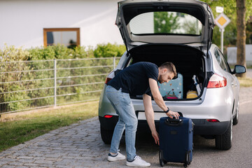 Handsome bearded man stand with suitcase in front of open car trunk. Travel road trip. Holiday summer vacation concept