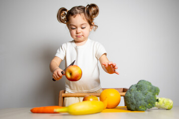 Fototapeta na wymiar Cute caucasian brunette toddler girl with two tails in white dress staying at the learning tower cutting apple on knife, Montessori method on white background 