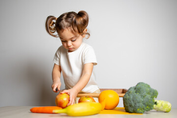 Fototapeta premium Cute caucasian brunette toddler girl with two tails in white dress staying at the learning tower and cutting apple, Montessori method on white background 