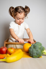 Fototapeta premium Cute caucasian brunette toddler girl with two tails in white dress staying at the learning tower and cutting grapefruit, Montessori method on white background 