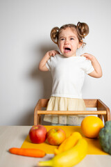 Cute caucasian brunette toddler girl with two tails in white dress staying at the learning tower crying, near vegetables, Montessori method on white background 