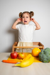 Fototapeta premium Cute caucasian brunette toddler girl with two tails in white dress staying at the learning tower crying, near vegetables, Montessori method on white background 