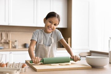 Happy beautiful baker kid girl in household apron preparing bakery meal in kitchen, cooking pastry...