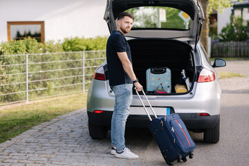 Handsome bearded man stand with suitcase in front of open car trunk. Travel road trip. Holiday...