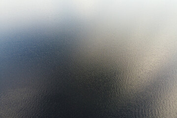 The tranquil expanse of a blue lake stretches endlessly in this top-down drone shot, where the subtle play of light and water coalesces into a soothing, monochromatic palette