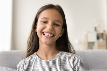 Happy cute pre teen kid girl looking at camera with toothy smile, talking on online video call, chatting on Internet, enjoying distant communication. Home head shot portrait of cheerful child