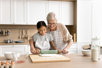 Happy grandmother in apron teaching cheerful granddaughter to bake homemade pastry food, helping to...