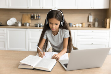 Focused pre teen schoolkid girl in wireless headphones studying from home, using laptop, listening video training class, writing notes in copybook, standing at kitchen table