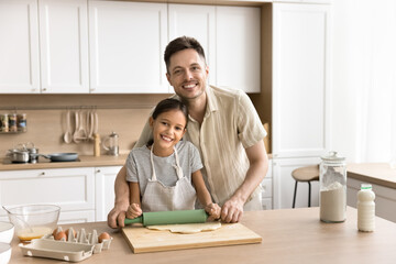 Cheerful preteen kid girl and happy young dad baking pastry food in home kitchen, rolling raw fresh dough on table, together, enjoying culinary hobby, looking at camera, smiling