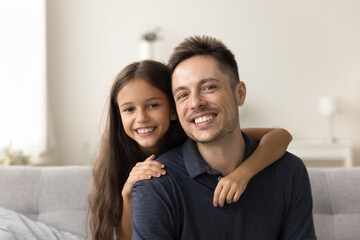 Happy handsome dad and cheerful tween kid girl looking at camera with toothy smiles, posing for home headshot portrait. Positive father piggibacking cute daughter head shot