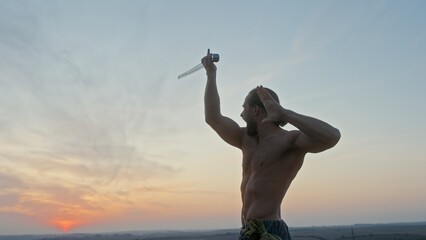 Confident strong man training with sword in nature during sunset