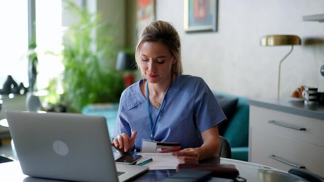 Nurse paying bills online, sitting in living room, working on notebook. Doctor in scrubs dealing with work issues from home, after workday, telemedicine consultation.