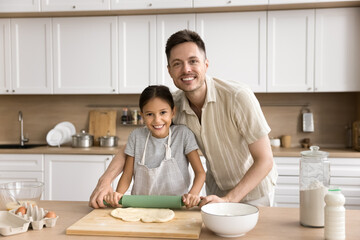 Obraz na płótnie Canvas Happy cooking blogger dad teaching cute kid girl in apron to bake homemade pastry meal, dessert, rolling dough for pitta, looking at camera, smiling for portrait, laughing, having fun
