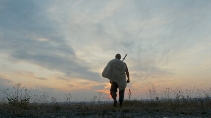 Back view of male barbarian with sword walking in meadow towards cloudy sundown sky