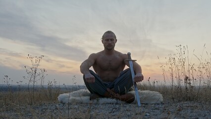 Muscular shirtless swordsman sitting cross-legged on a sheepskin spread out on the ground meditating at sunset with his sword resting against his knee in a rotating panning view at ground level