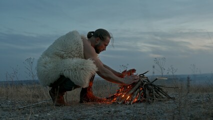 Man in animal hide kneeling and blowing at burning campfire against cloudy sky in evening in wilderness