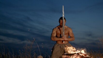 Shirtless warrior putting sword into ground near fire