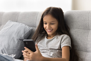 Happy beautiful pre teen girl kid browsing social media on smartphone, chatting online, playing virtual game on Internet, resting on sofa, enjoying leisure, online communication