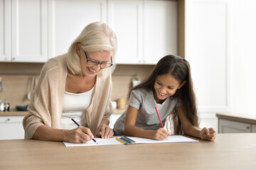 Cheerful grandmother and pretty grandchild girl drawing creative doodles in paper album together, sitting at kitchen table, discussing art, creativity, hobby, school homework, laughing