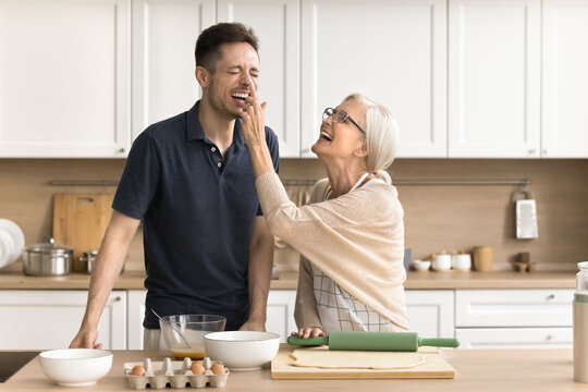 Joyful older mom and son baking together, cooking breakfast in kitchen, having fun, laughing, enjoying family culinary hobby. Cheerful senior mother touching face of adult child with floury finger - Powered by Adobe