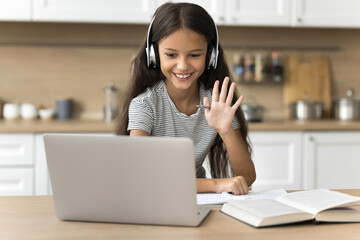 Cheerful preteen school girl in wireless headphones talking on video conference call on laptop, attending distant class, lesson, studying at home, doing homework task, waving hello at computer