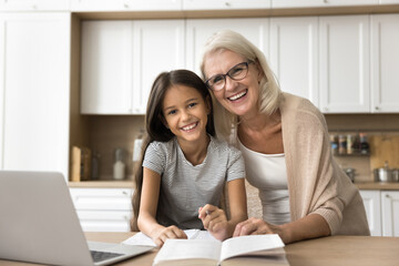 Cheerful pretty grandmother and happy cute granddaughter schoolchild doing school homework task, sitting at kitchen table with textbook and laptop, looking at camera with toothy smile for portrait