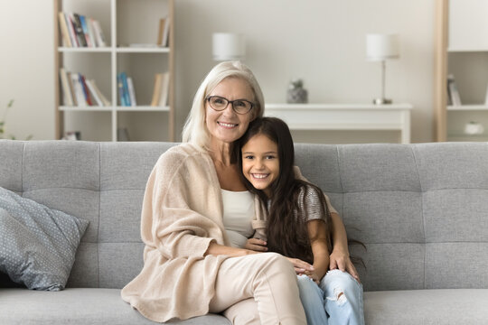 Happy positive loving granny and cute granddaughter girl hugging with love, affection on comfortable couch, looking at camera with toothy smile for family portrait, enjoying homey leisure, meeting
