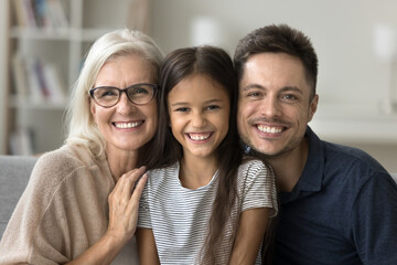 Happy cute little girl, young dad and grandmother sitting close on sofa, hugging, looking at camera, laughing, enjoying family leisure at home. Daughter kid, father and grandma head shot portrait