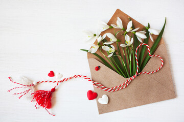 Envelope with spring flowers snowdrops and red and white martenitsa, tassel cord, a symbol of the Martisor holiday, Baba Marta on a white wooden background. Greeting card. - 732743714