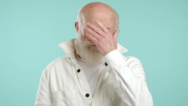 A bashful elderly man in a youthful white jacket covers his face with his hand against a turquoise backdrop, portraying a moment of shyness or embarrassment. Camera 8K RAW. 