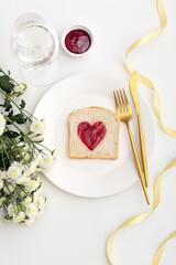 Sandwich with jam in the shape of a heart on a white plate and with gold kitchen utensils. Flowers and a glass of champagne on a white table. Top view.